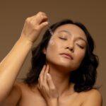 Asian woman with closed eyes applying skin tonic to her cheek with dropper cap. Femininity, body, skin and beauty treatments, unaltered.